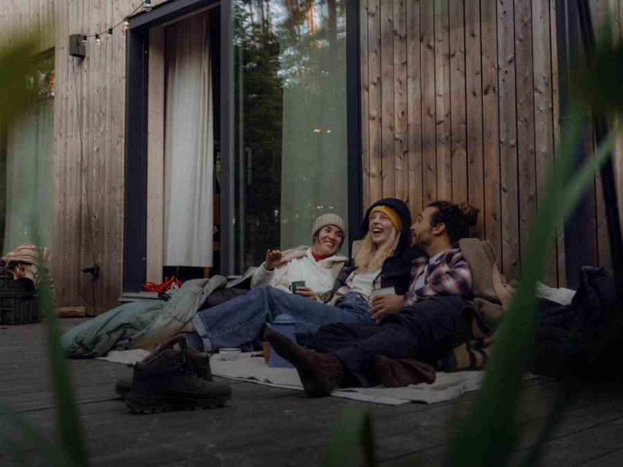 Tre persone in abiti casual da esterno siedono accoccolate insieme sulla veranda di una capanna di legno nella foresta.