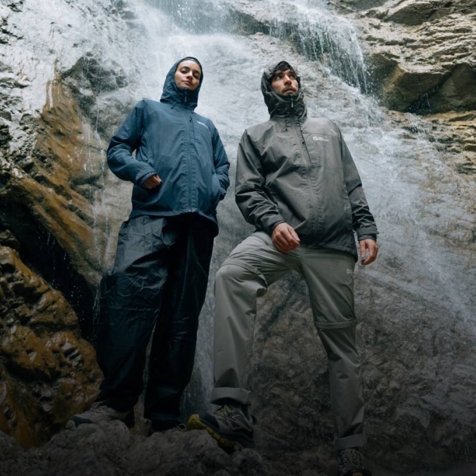 Zwei Wanderer in sportlichen Regenjacken stehen vor einem Wasserfall